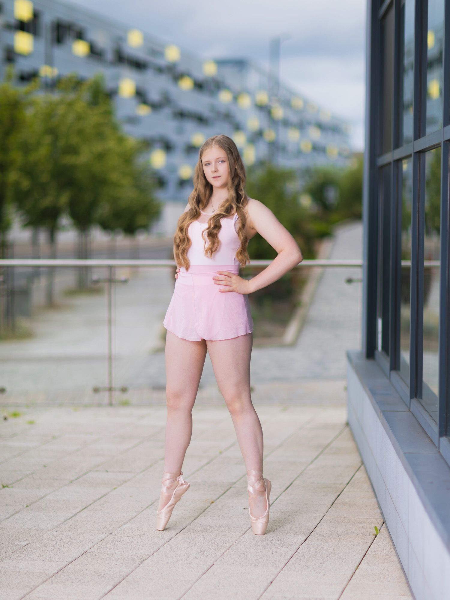 Young ballerina in pink tutu and pointe shoes poses gracefully outdoors near modern building.
