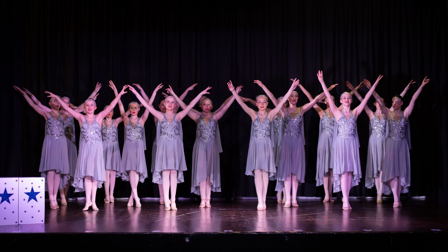 Ballet dancers in gray dresses perform on stage with arms raised, lined up in a graceful synchronized routine.