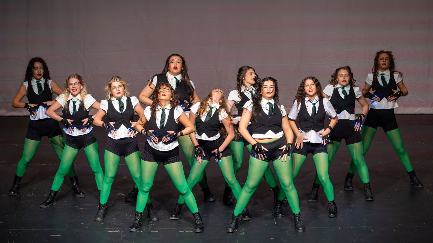 Dancers in green tights, black skirts, and vests perform energetically on stage.