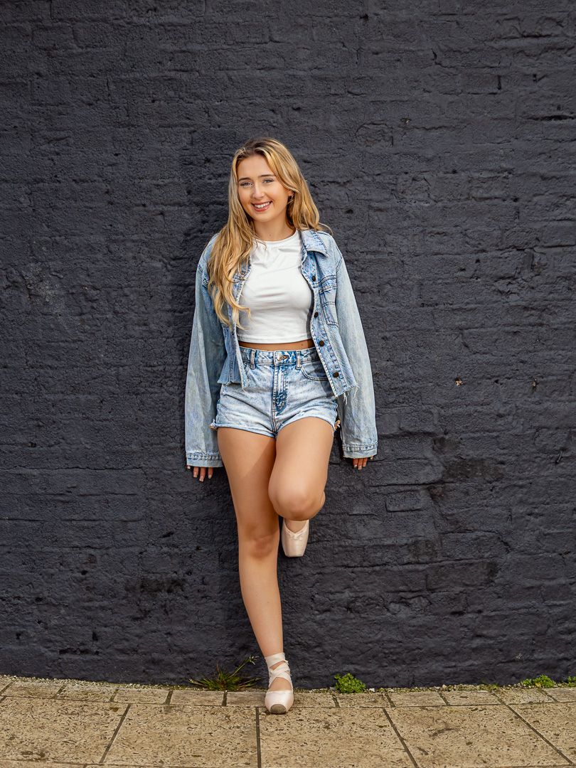 Street style poses against black brick wall in cropped denim jacket and shorts.