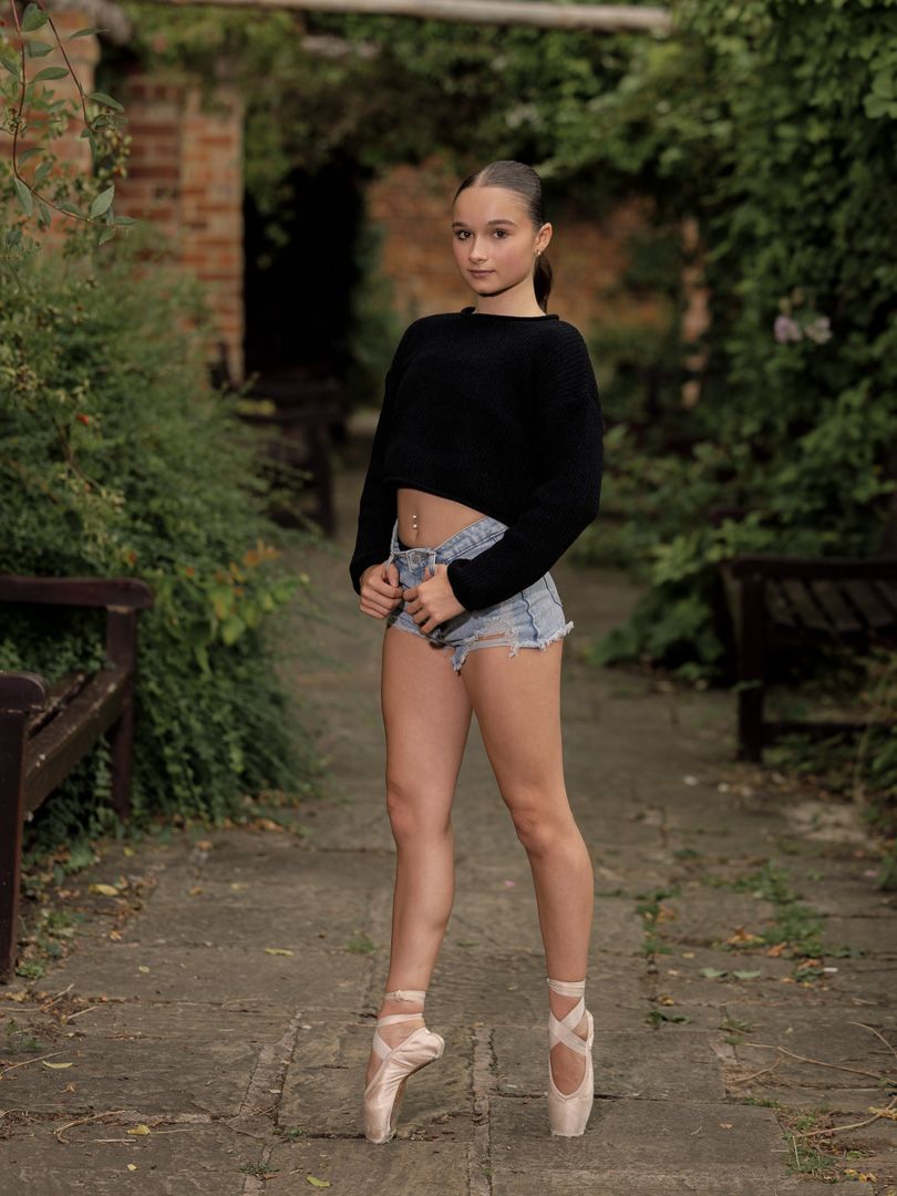 Dancer in black top and denim shorts practices ballet poses on outdoor pathway.