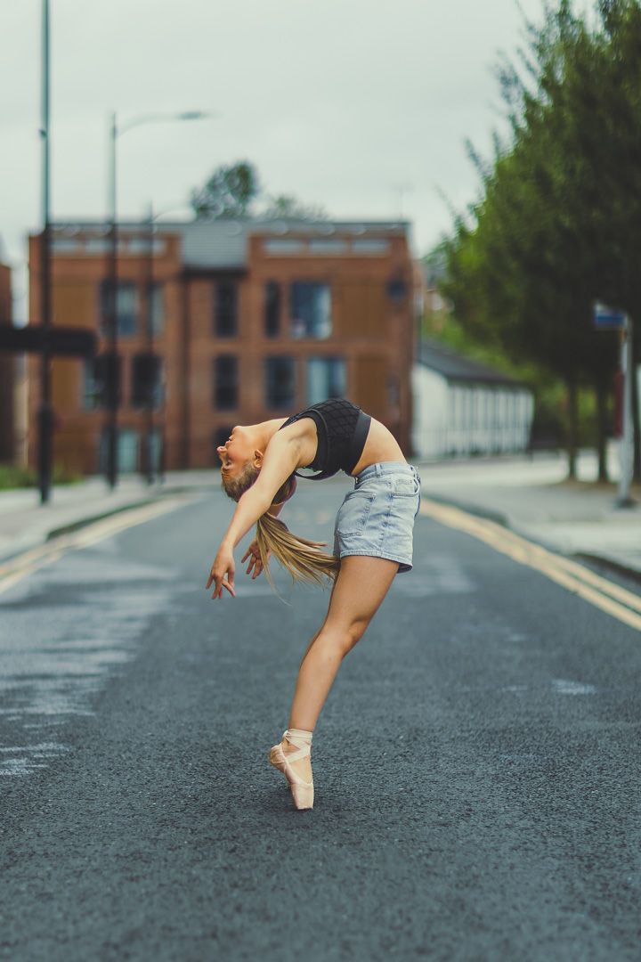 Ballerina dancing en pointe on an urban street in denim shorts and black top, arching backwards gracefully.
