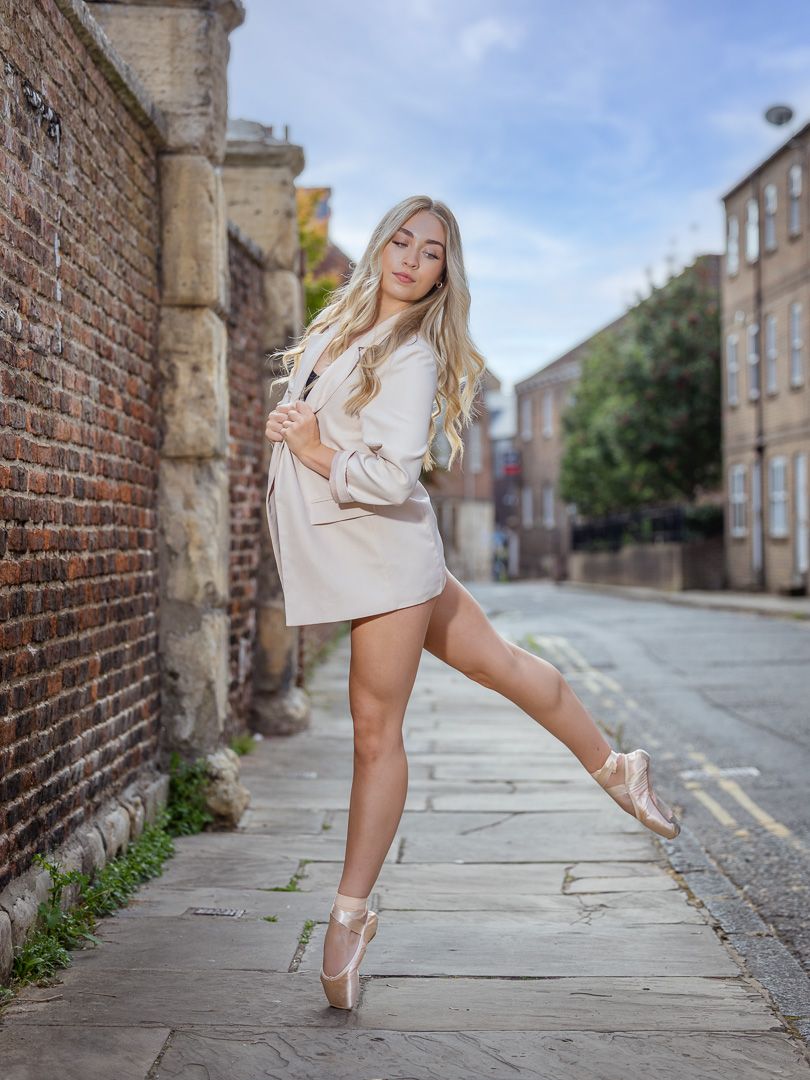 Blonde ballerina in cream outfit poses en pointe on cobblestone street beside brick wall in urban setting.