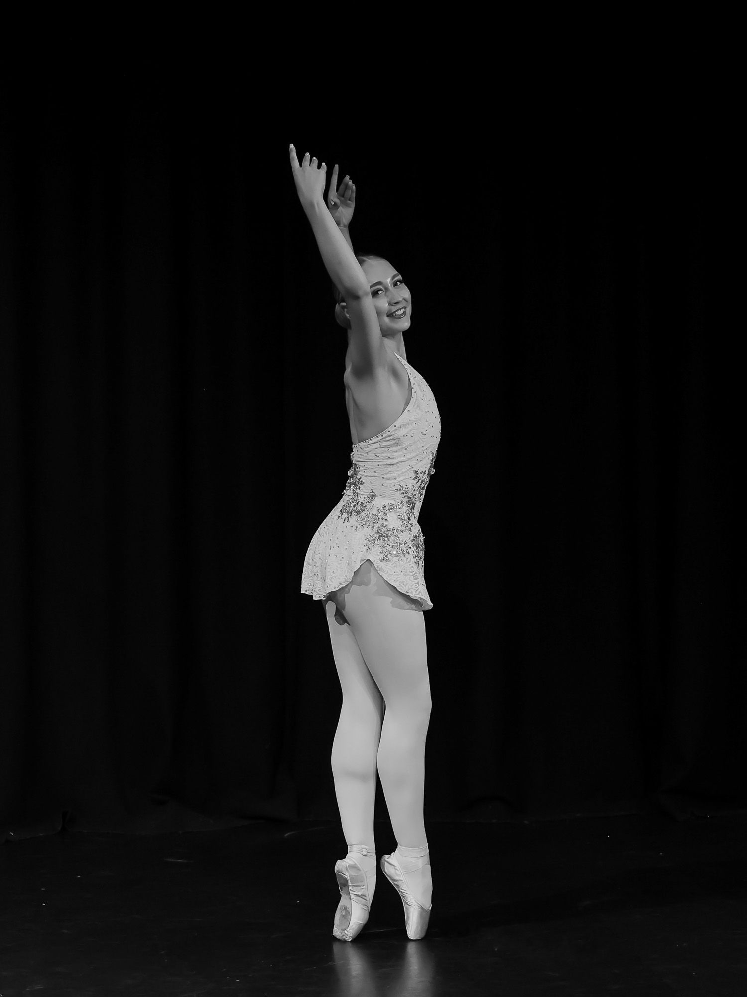 Black and white photo of a smiling ballerina in white dress on pointe with arm raised on stage.