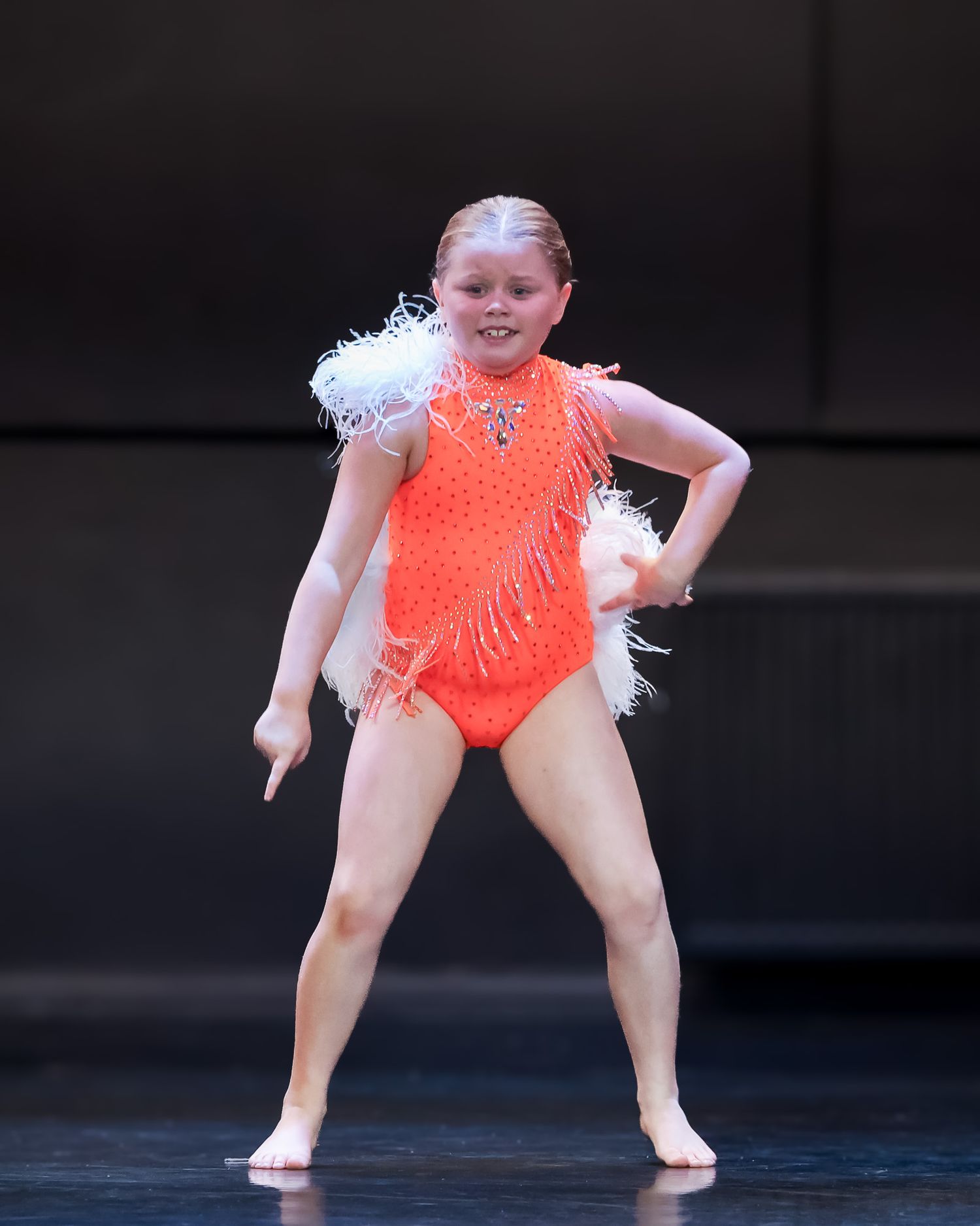 Young girl performing on stage in orange sequined dance costume with white feather trim under bright spotlights.