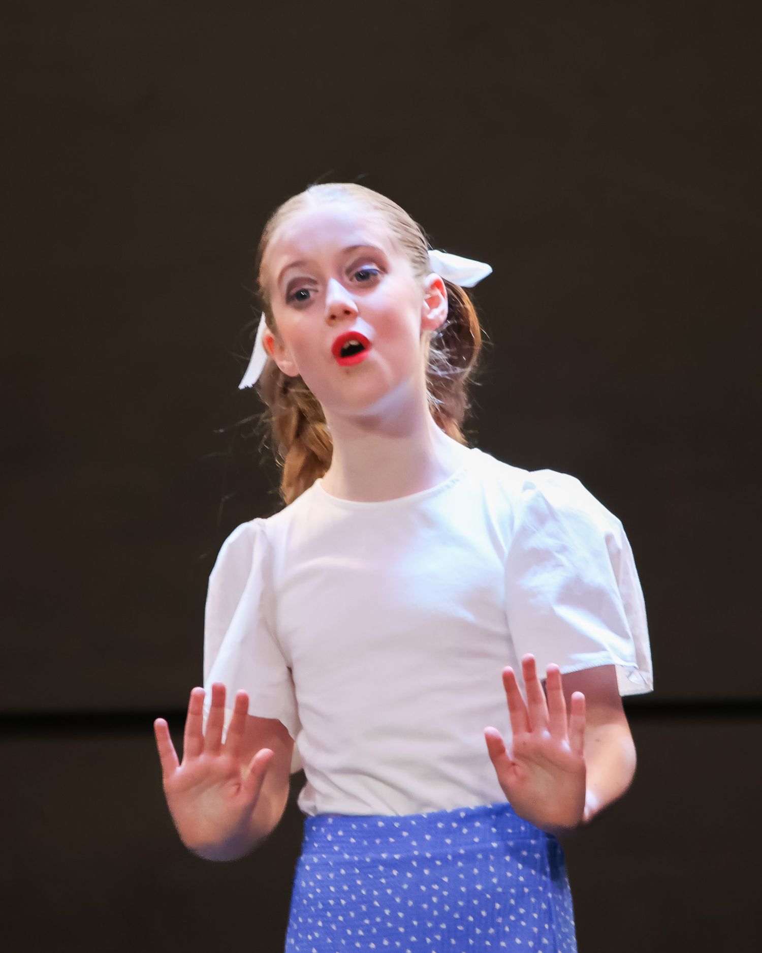 Young girl in white top and blue polka dot skirt performing on stage with hands raised.