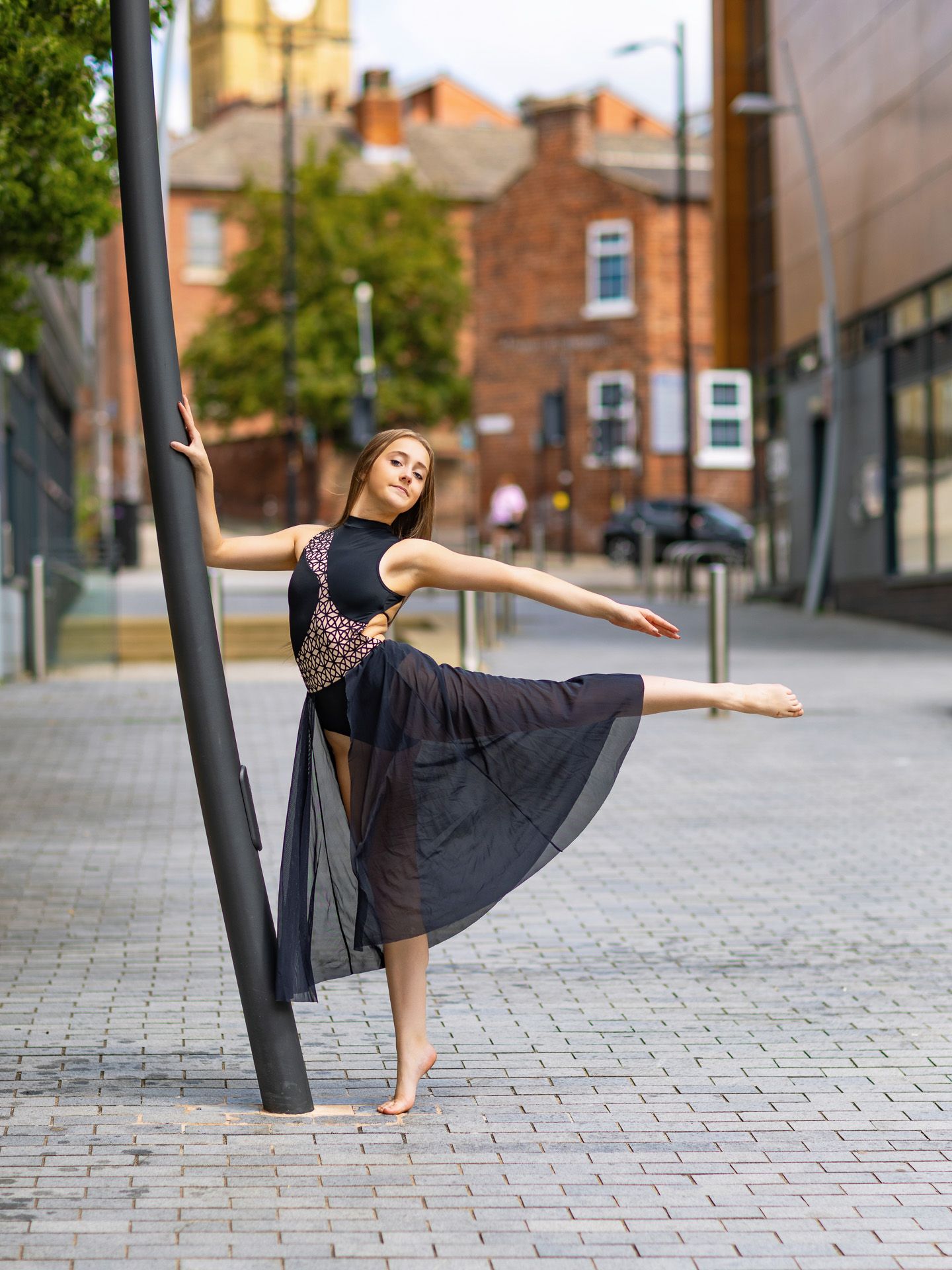 A graceful dancer in a black skirt poses barefoot beside a street pole in an urban setting.