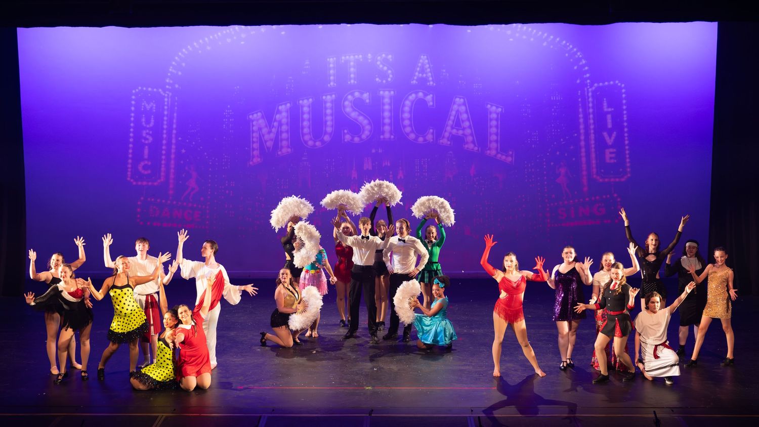 Colorful musical theater cast poses on stage under 'It's A Musical' sign with feathered costumes and bright lighting.
