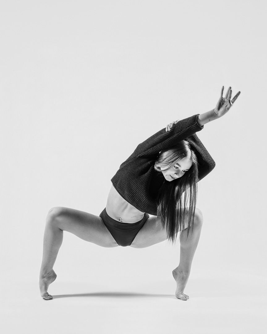Contemporary dancer in black attire performs expressive pose with bent knee and raised arm against white backdrop.