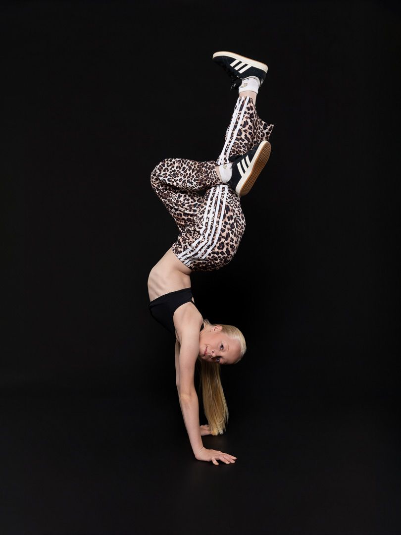 Young blonde gymnast doing a handstand in leopard print leggings and Adidas shoes on black background.