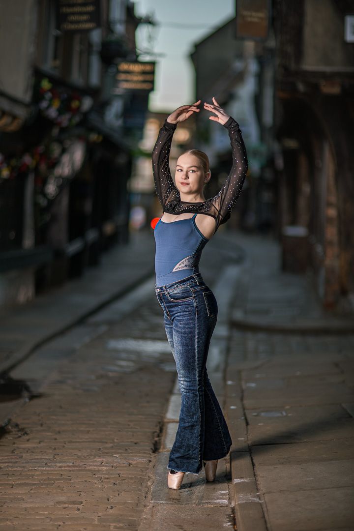 Ballet dancer in blue crop top and jeans strikes graceful poses on cobblestone street.