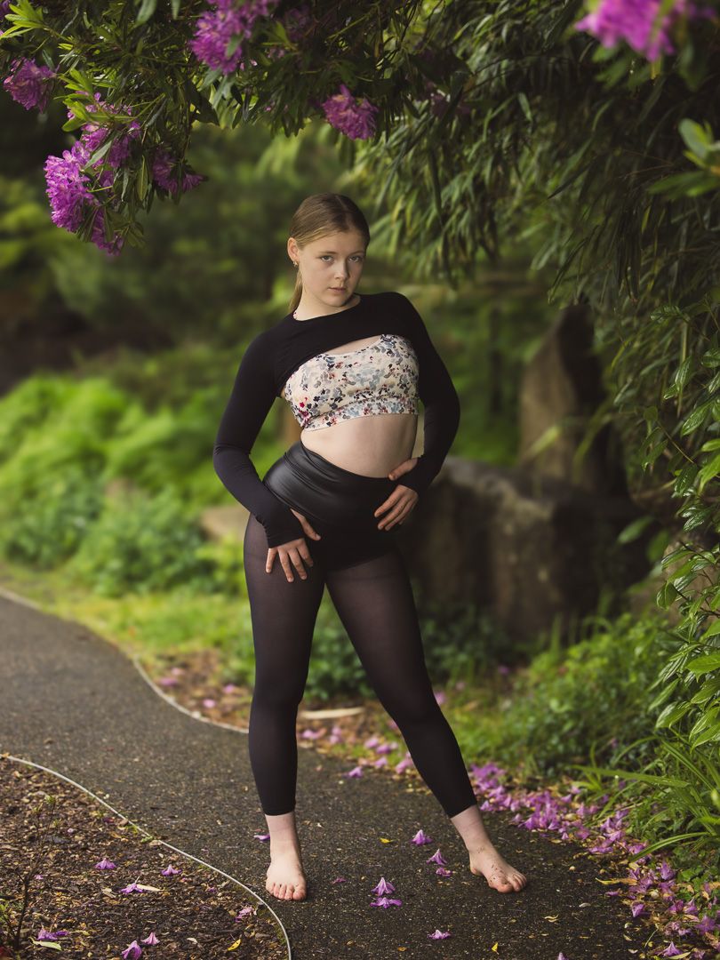 Contemporary dancer poses on path beneath blooming purple flowers in athletic wear.