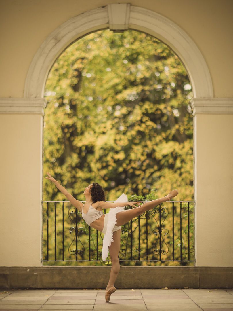 Ballet dancer performs arabesque pose under elegant arched architecture with natural lighting.