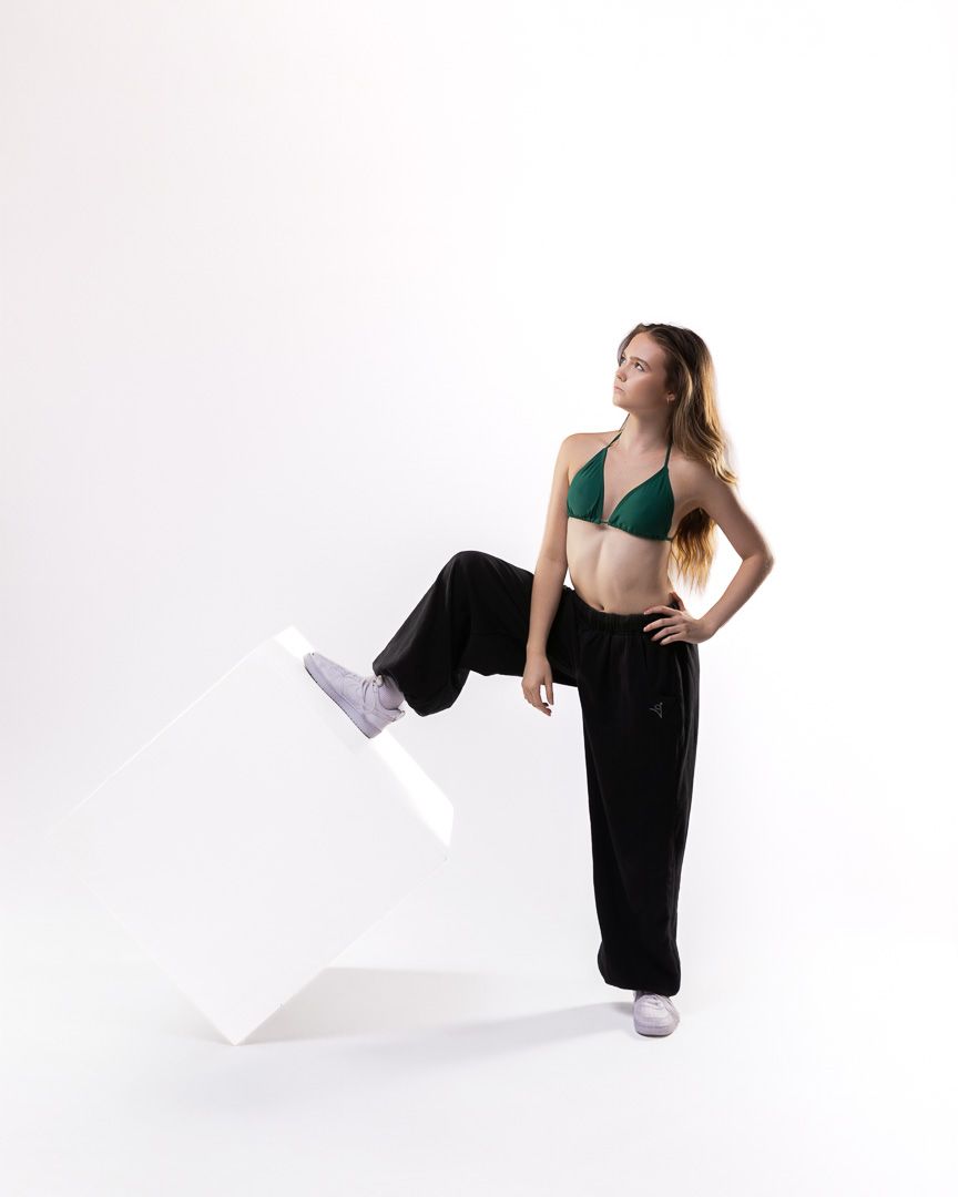 Woman in green bikini top and black wide-leg pants poses beside a white geometric cube in studio.