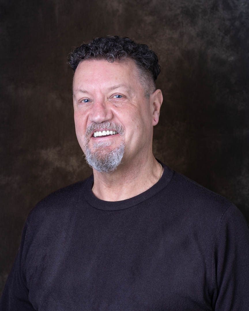 Middle-aged man with dark curly hair and goatee smiles in a casual black shirt for a professional portrait.
