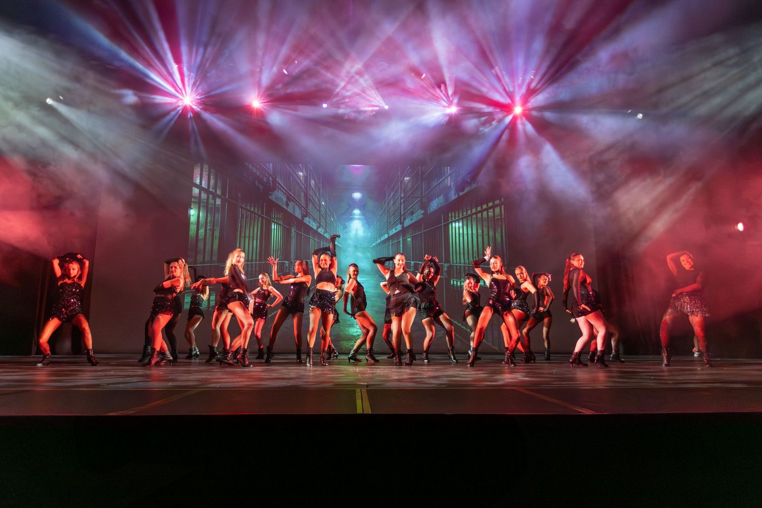 Dancers perform on a dramatic stage with red and teal spotlights during a high-energy group dance recital.