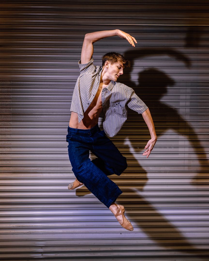 Male ballet dancer leaping mid-air in striped shirt and blue pants, casting dramatic shadow on metal shutter backdrop.