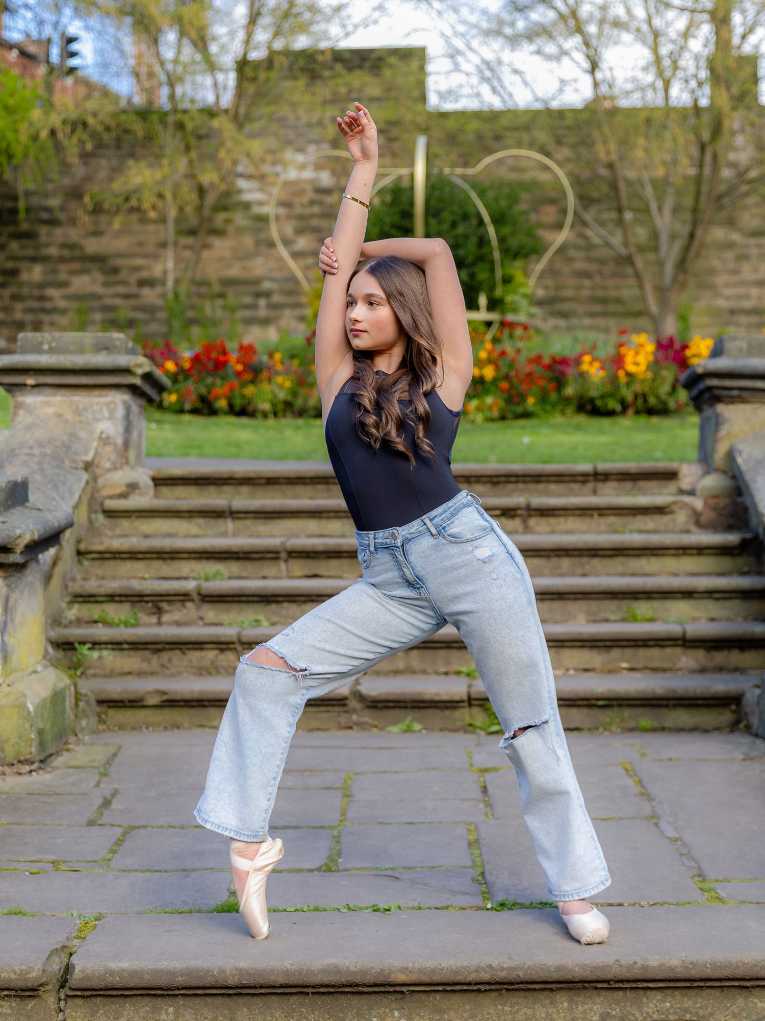 A young dancer poses in ripped jeans and ballet pointe shoes on stone steps surrounded by colorful flowers.