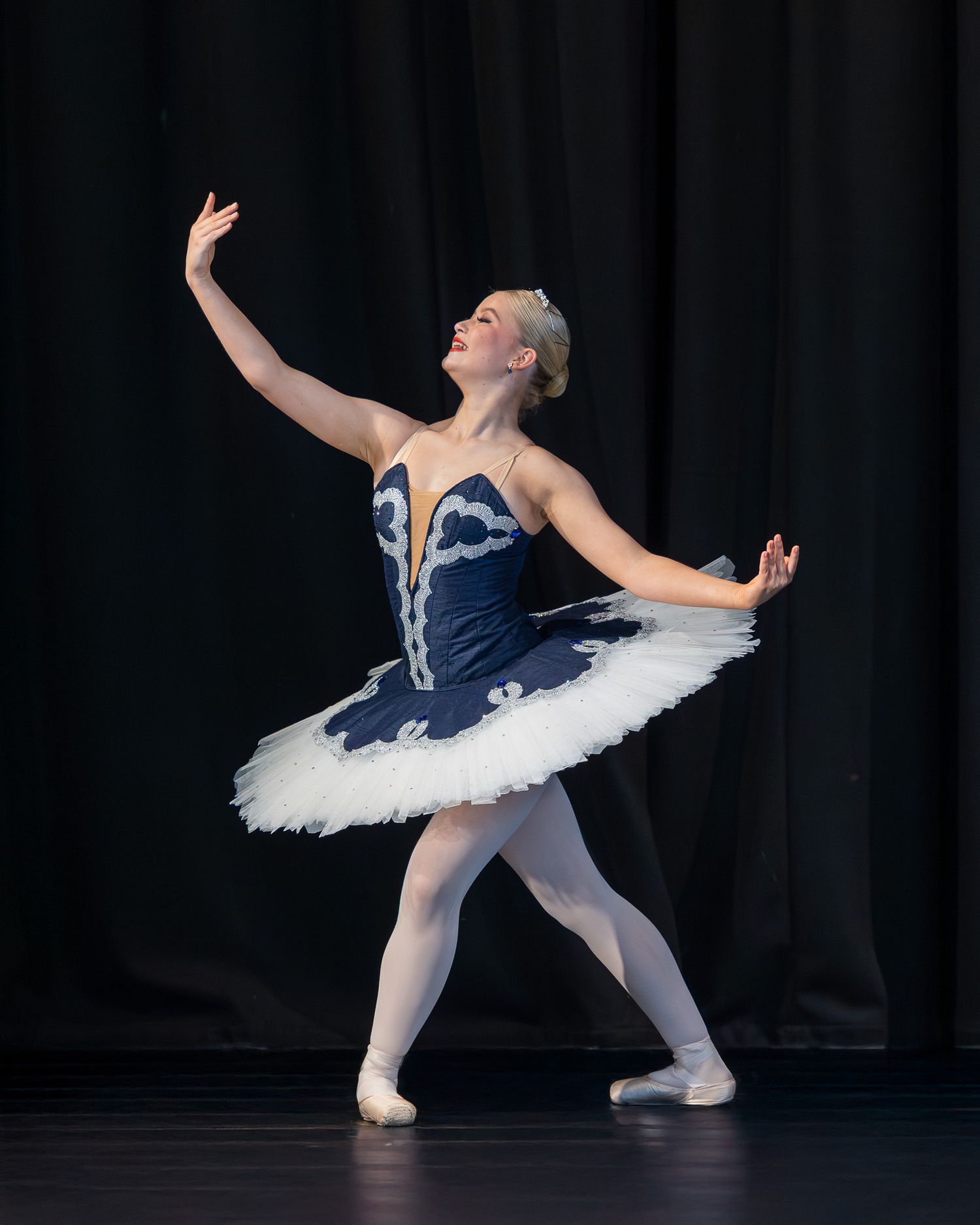 A young ballerina performs in a navy blue tutu and white tights on stage during a classical ballet recital.