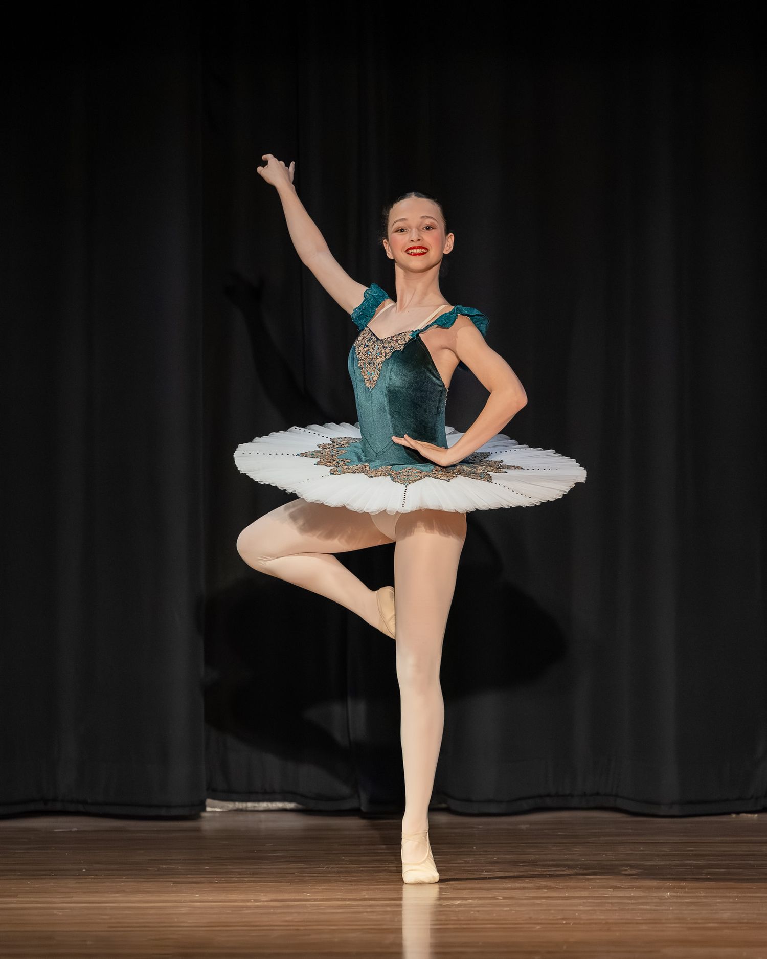 Young ballerina in teal velvet tutu performs elegant pose on stage under spotlight during ballet recital.