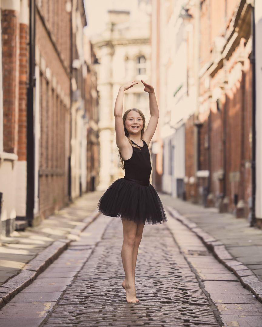 Ballet dancer performs graceful pose on cobblestone street in black tutu dress.