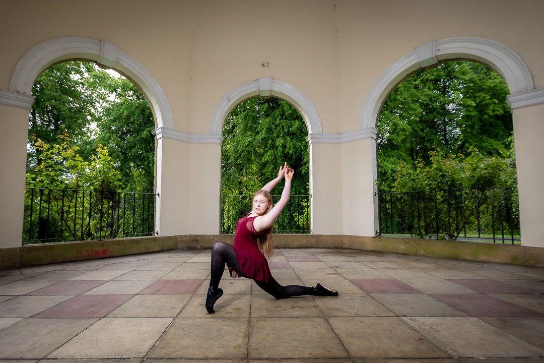 Dancer in burgundy outfit performs split leap under classical arched colonnade.