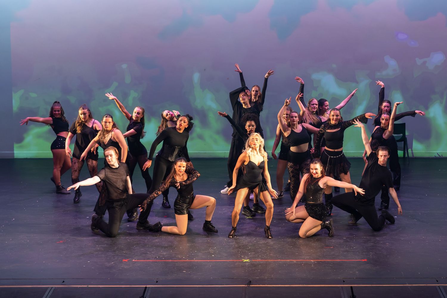 Dance ensemble in black costumes poses dramatically on stage with colorful backdrop during a theatrical performance.