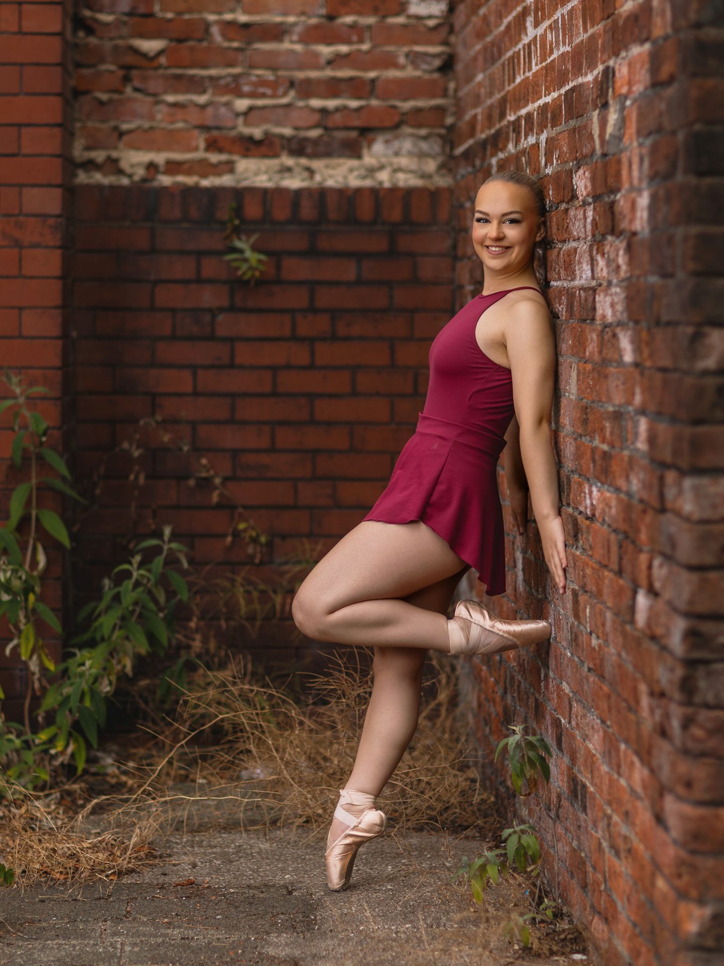 A ballet dancer in a burgundy leotard poses on pointe shoes against a rustic brick wall outdoors.