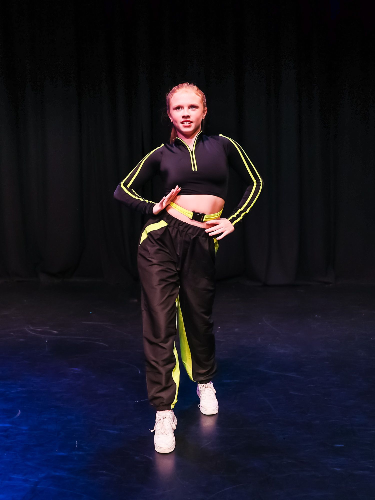 Young dancer poses on stage in black and neon yellow crop top and joggers with white sneakers under blue stage lighting.