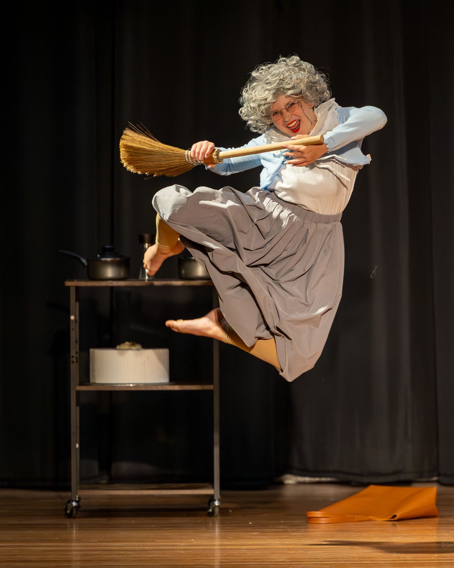 Performer dressed as a witch leaps mid-air holding a broom on stage, wearing gray wig and costume.