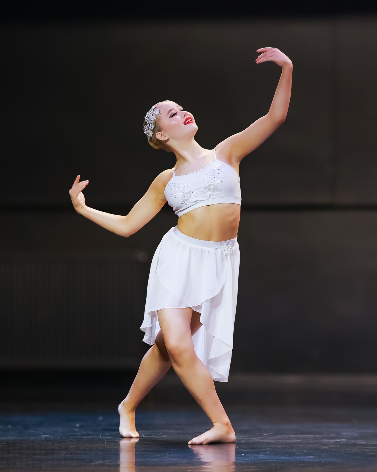 Young dancer in white crop top and skirt performs graceful contemporary dance pose on stage.