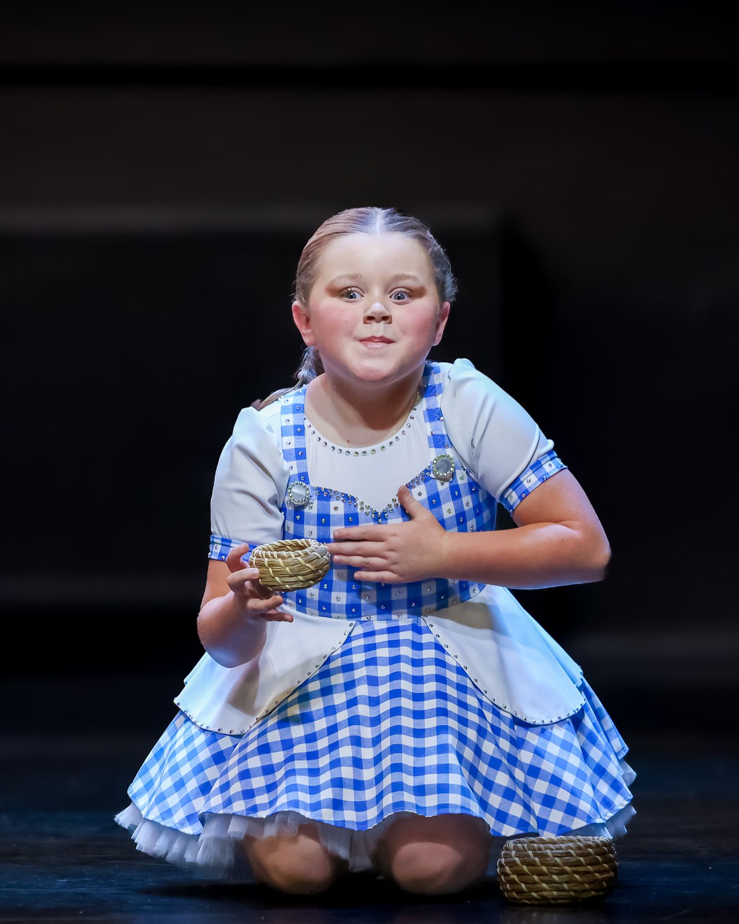 Young girl dressed as Dorothy from Wizard of Oz in blue gingham dress, holding basket on stage.