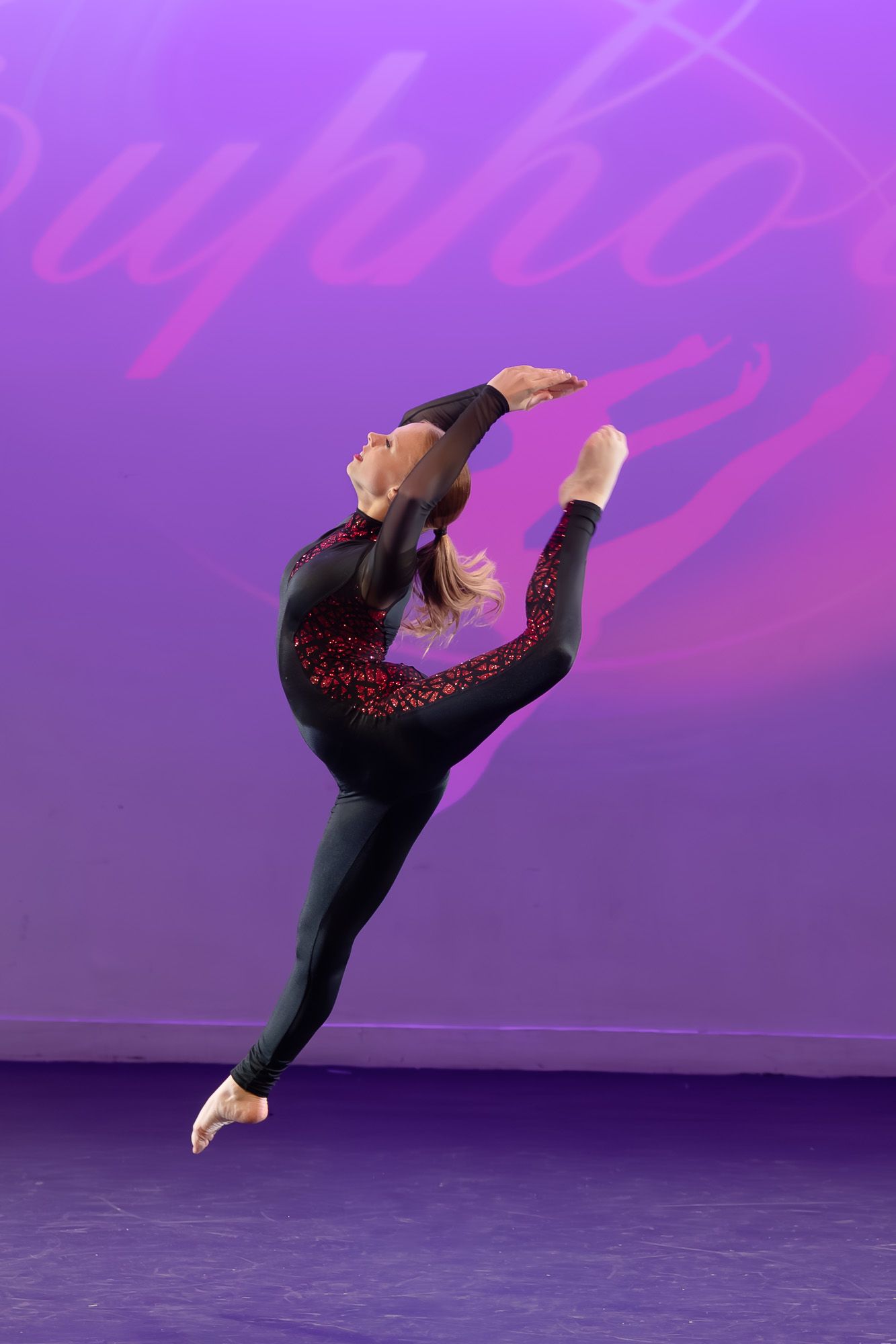 A gymnast in a black and red sequined leotard performs a graceful leap against a purple stage backdrop.