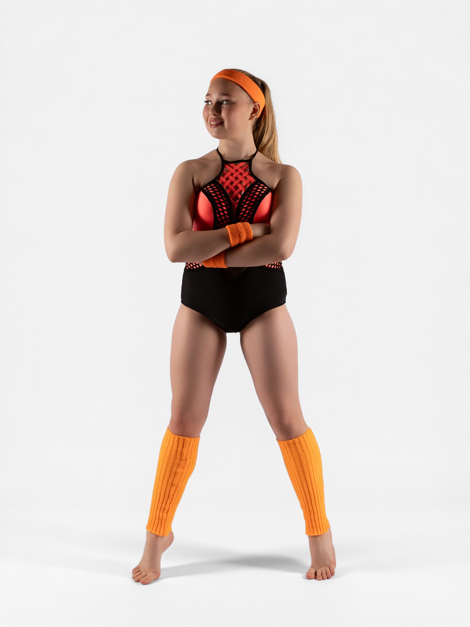 Young female dancer in black bodysuit, orange leg warmers, headband, and wristbands poses confidently with arms crossed.