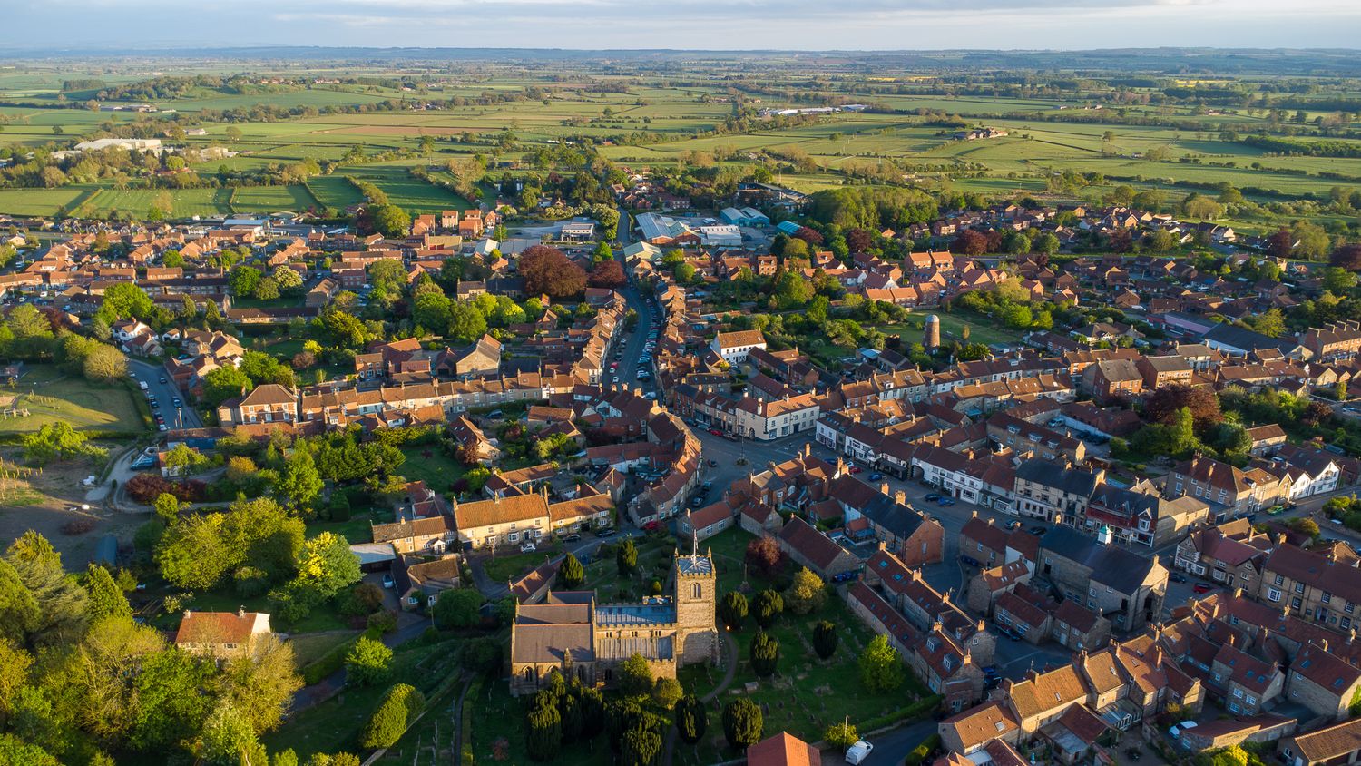 Aerial view of a quaint English village with red-roofed buildings nestled in rolling green countryside.