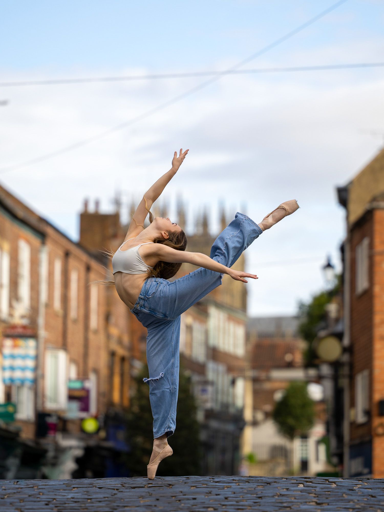 A ballet dancer performs an arabesque in jeans on a cobblestone street with brick buildings in the background.