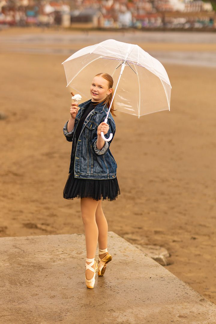 Young girl in denim jacket and ballet shoes holds white umbrella and ice cream cone on sandy beach.