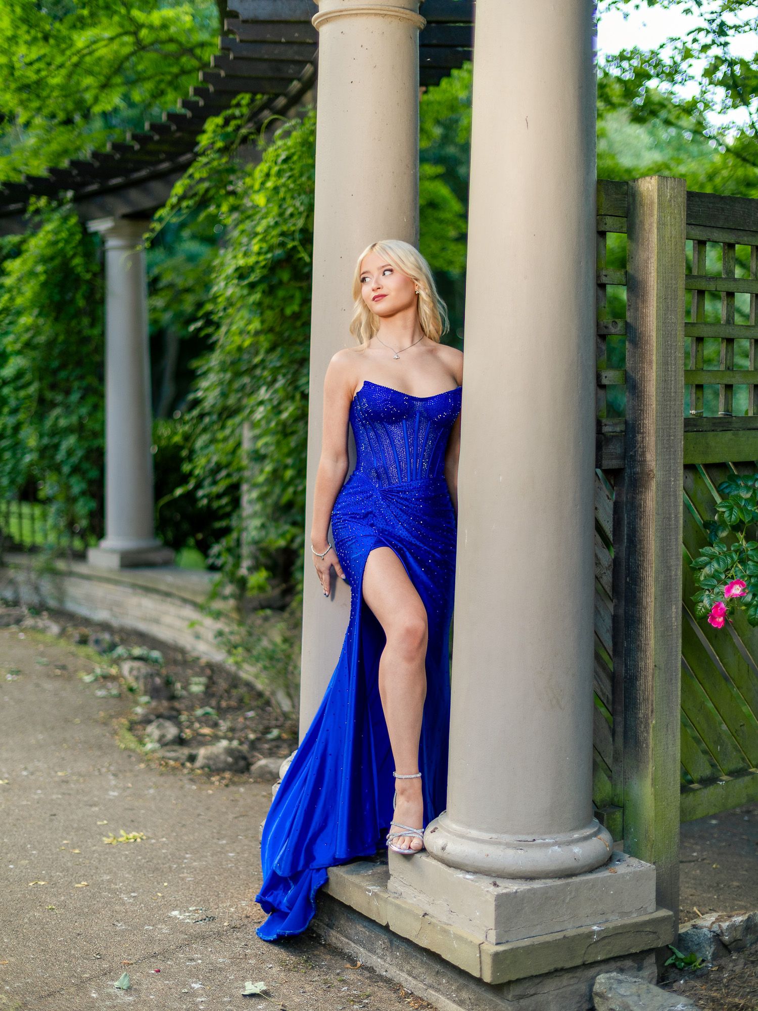 Woman in royal blue satin corset gown with high slit poses between stone columns in lush garden setting.