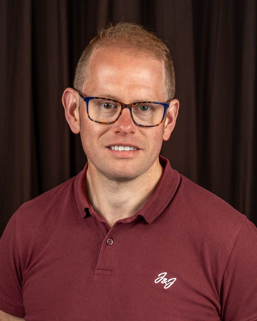 Man with blue glasses and maroon polo shirt smiling in a professional headshot photo against dark background.
