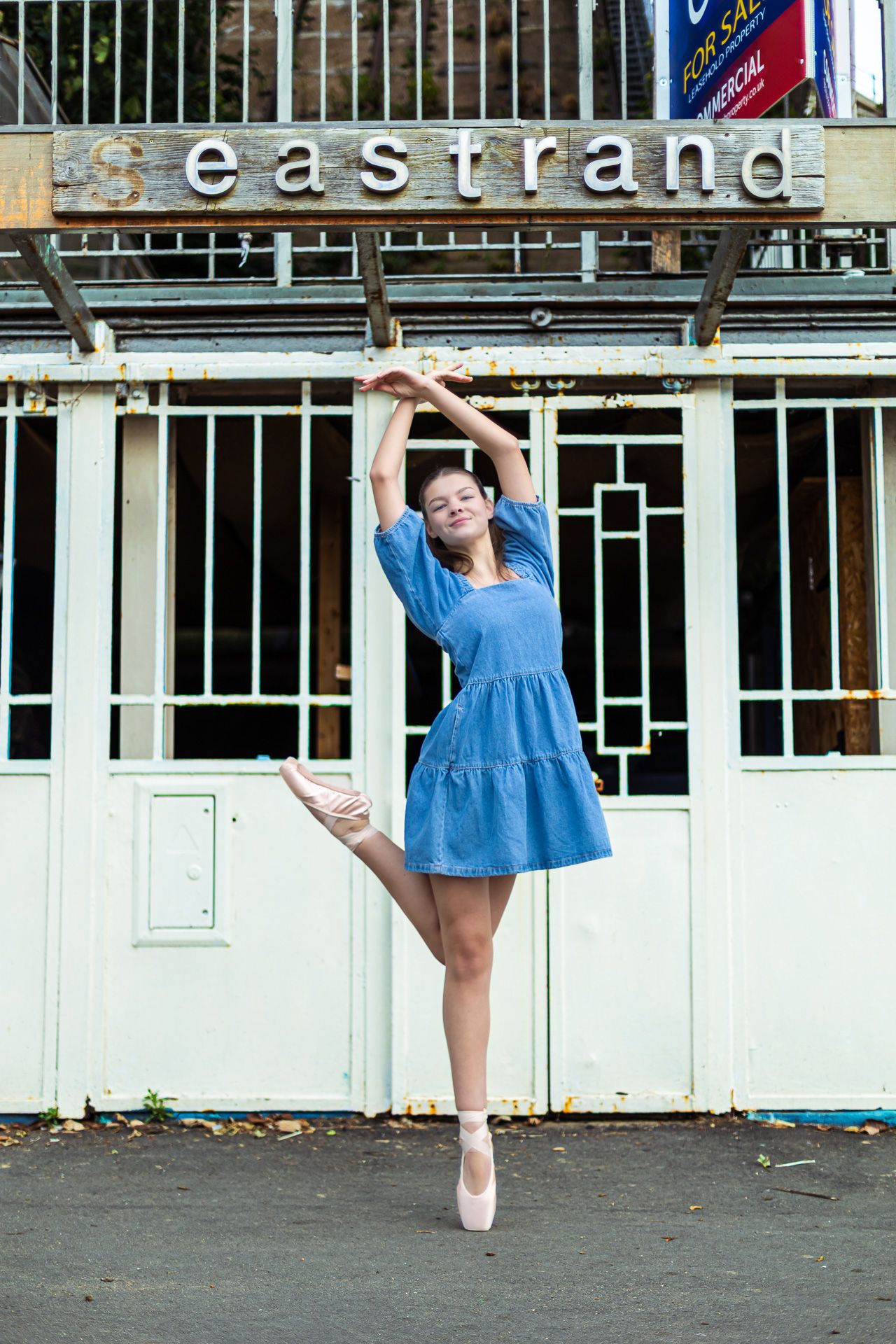 Ballerina in blue dress and pointe shoes performs elegant leg extension pose outside Eastrand building.