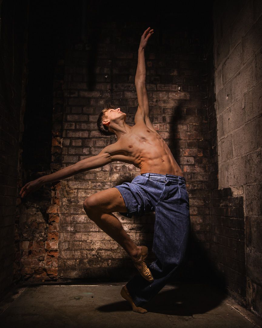 Shirtless male dancer poses dramatically against a rustic brick wall, wearing blue shorts and gold shoes.