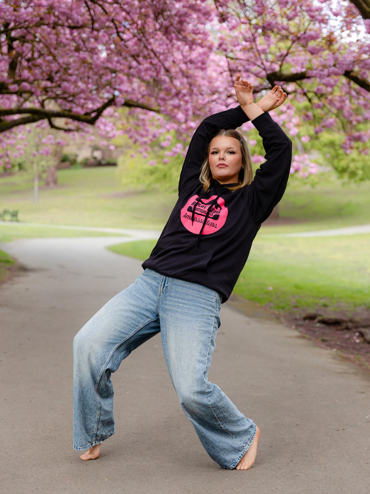 Woman in black graphic sweatshirt and wide-leg jeans poses barefoot under pink cherry blossom trees in a park.