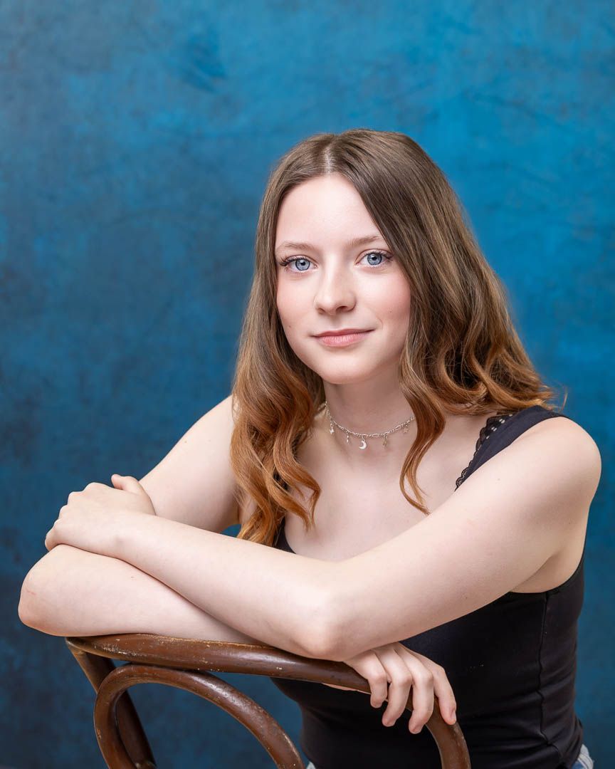 Young woman with auburn hair posing gracefully on a wooden chair against a blue textured studio background.