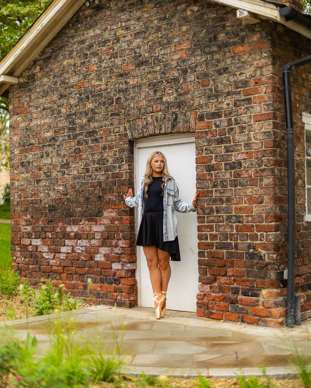 Blonde woman in black dress, denim jacket, and ballet shoes posing by white door of rustic brick building outdoors.