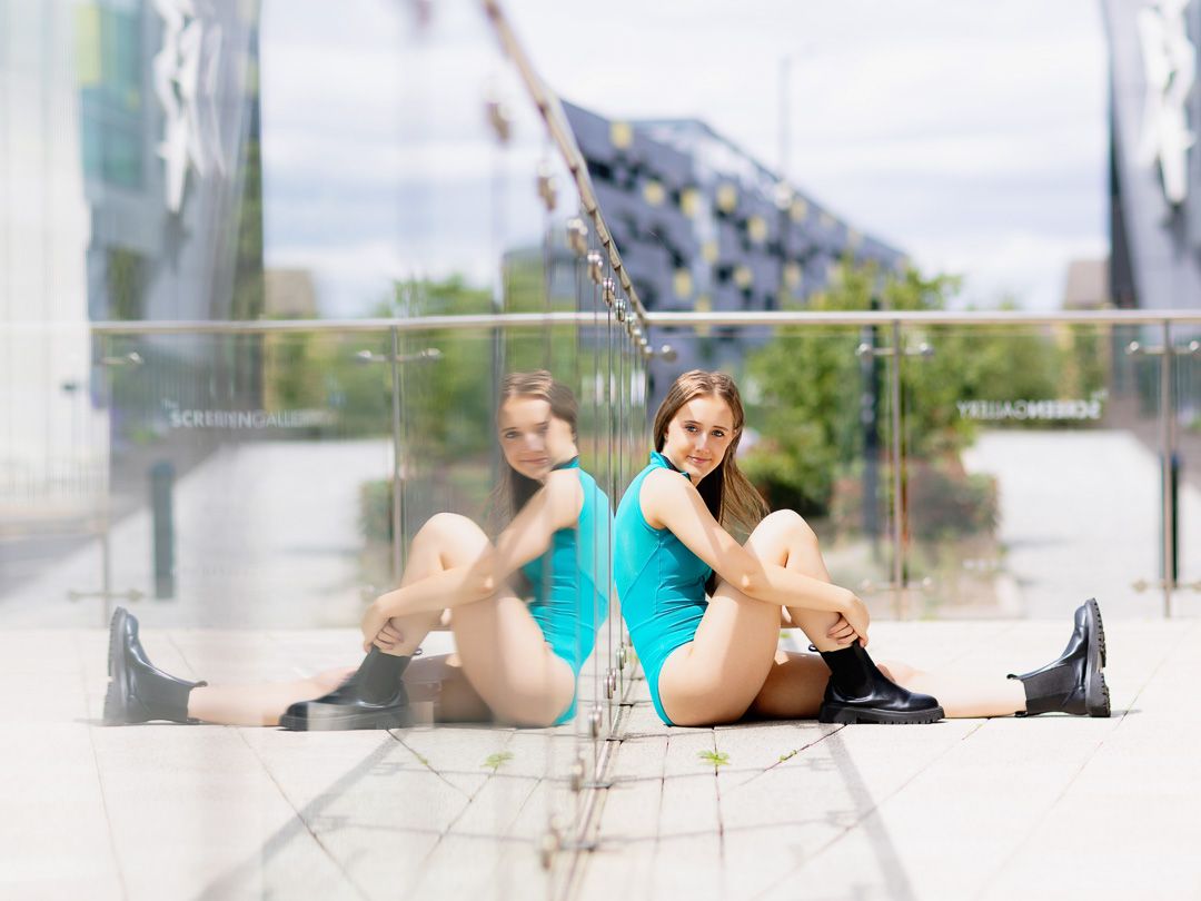 Two flexible young women in teal outfits sit back-to-back on an urban plaza floor with a glass wall reflection.