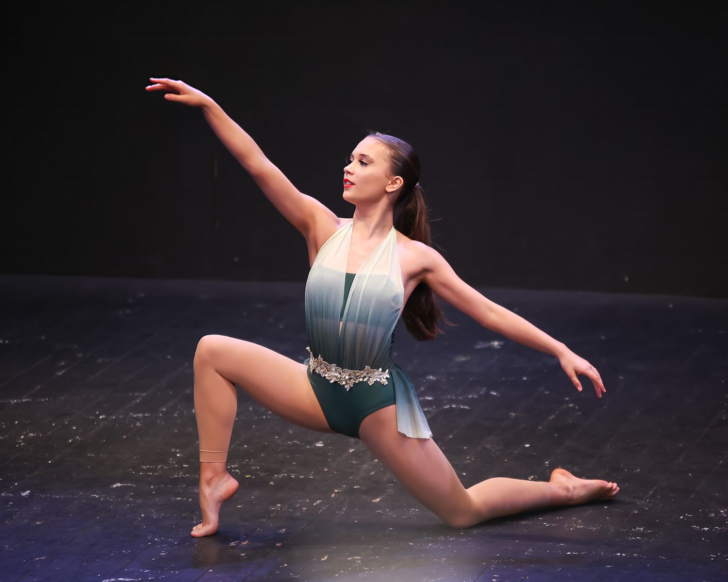 Young female dancer performs a graceful lunge pose in a teal ombre leotard on a dark stage.
