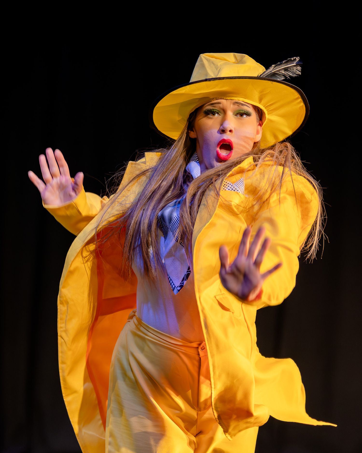 Performer in bright yellow suit and hat with feather gestures dramatically on stage with open mouth and raised hands.