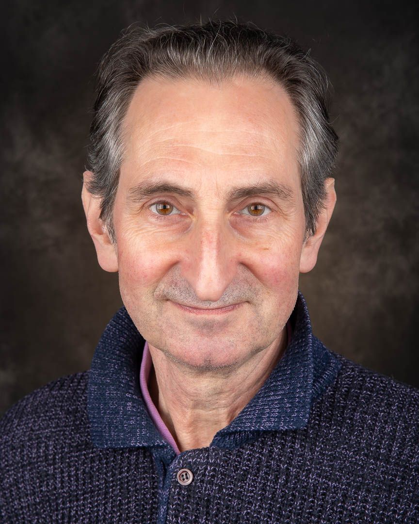 Older man with gray hair smiling warmly, wearing a blue knit sweater, professional headshot.