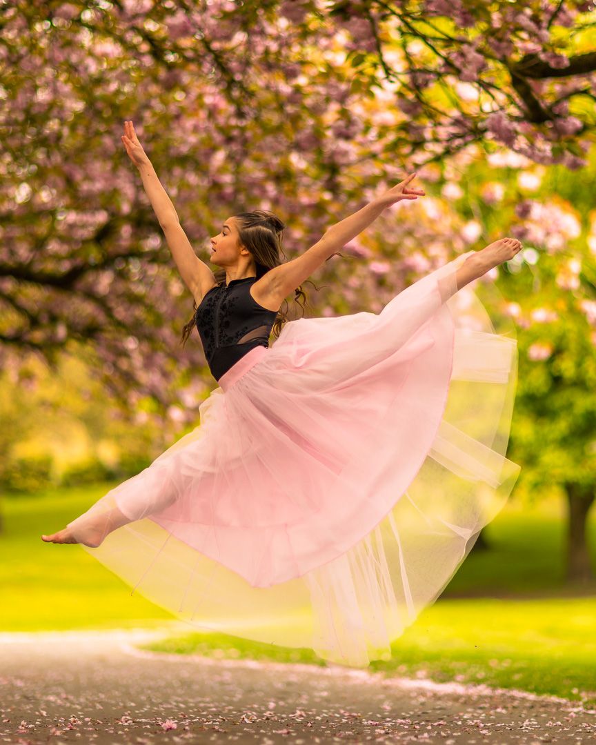 Ballerina leaps gracefully in pink tulle skirt among cherry blossoms in a lush green park.