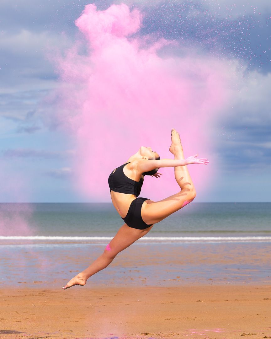 Dancer leaping on beach in black bikini with pink powder cloud explosion against cloudy sky backdrop.